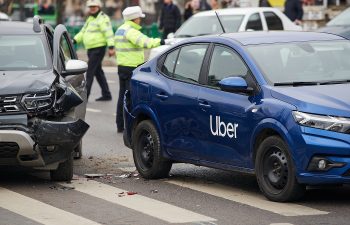 Car accident involving a blue Uber vehicle and a black vehicle on a street. Police officers are on the scene, with other cars and pedestrians visible in the background.